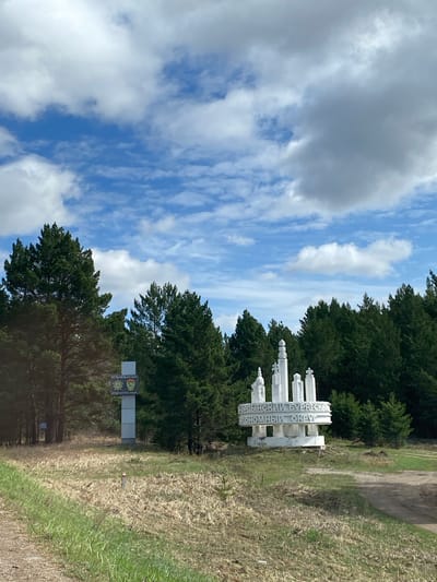 The image depicts a landscape featuring a combination of natural and man-made elements. A white monument with Cyrillic script stands prominently, possibly indicating the entrance to the Ust-Ordynsky Buryat Okrug in Russia. The text on the monument includes "УРДЫНСКИЙ БУРЯТСКИЙ НОННЫЙ ОКРУГ."

Adjacent to the monument, there is a sign marked with a coat of arms or emblem along with additional Cyrillic text. The scene includes a backdrop of trees, grassy areas, and a sky with a mix of blue and clouds. The photograph appears to have been taken during the day in Jerdovka, Russia, based on the sign and monuments shown in the scene.