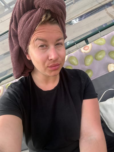 A woman with blonde hair wrapped in a dark brown towel is looking at the camera with her head tilted slightly. She is wearing a black t-shirt. The background consists of wooden planks, a green metal railing, and a fabric with an avocado print. A white bag is partially visible on the right side of the image. The scene appears to be outdoors, possibly on a balcony or patio. The overall lighting suggests daytime. The location is identified as Zamosh'e (Zaklinskoe S/P), Russia.