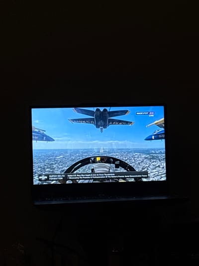 The image displays a television screen showing a video game simulation of fighter jets in a "Super Delta Formation" flying over a city. The screen is mostly filled with the game's graphics, depicting several U.S. Navy Blue Angels and U.S. Air Force Thunderbirds flying in formation against a blue sky. The perspective is from the cockpit of one of the aircraft, showing the dashboard and controls. Overlay text at the bottom of the screen reads "SUPER DELTA FORMATION" and "Unites U.S. Navy Blue Angels & U.S. Air Force Thunderbirds - Showcasing The Precision & Professionalism Of The Nation's Premier Aerial Demonstration Teams." In the upper right corner of the screen, the logo "MUCKS PORT 2HD LIVE" is visible. The television is positioned in a dark room, with only the screen illuminated. There is no indication of the city of Enschede, Netherlands, being present in the image.