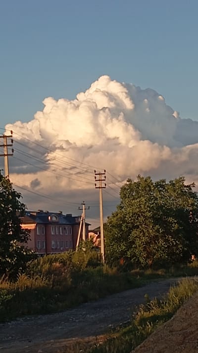 The image depicts a cloudy sky with a large, cumulonimbus cloud formation dominating the center. In the middle ground, a red brick building with multiple windows is visible, partially obscured by trees and vegetation. Several utility poles with wires run across the frame, suggesting an urban or suburban environment. The foreground shows a dirt road or path with grass and weeds growing along its edges. The scene appears to be set in the city of Baksan, Russia, likely during daylight hours given the lighting conditions. There are no people or animals readily apparent in the image. The overall atmosphere is calm and naturalistic, with a focus on the atmospheric cloudscape and the surrounding landscape.