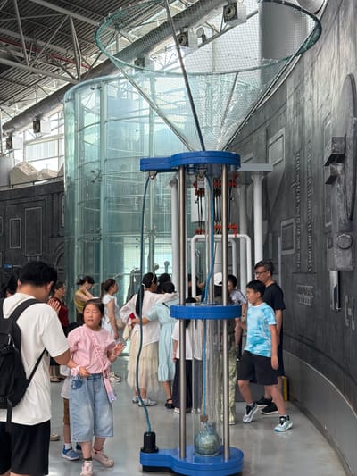 In an indoor exhibition space within Jiangbeicheng Subdistrict, China, a group of people, including adults and children, are gathered around a large, blue, metal and plastic exhibit. The exhibit features a glass tube in the center, connected to tubes and pulleys at the top and bottom. A green net structure extends upwards from the top of the exhibit. To the left of the exhibit is a tall, cylindrical glass structure, possibly part of another exhibit or architectural element, behind which is a large window showing a bright, overcast sky. The background consists of a dark gray, textured wall with various rectangular and circular indentations, resembling panels. The floor is a smooth, light gray material. Several individuals are looking at the exhibit or interacting with each other. A young girl in a pink shirt and denim shorts is in the foreground, looking towards the exhibit and gesturing with her hands. A young boy in a blue tie-dye shirt and black shorts is standing to the right of the exhibit, also looking at it.