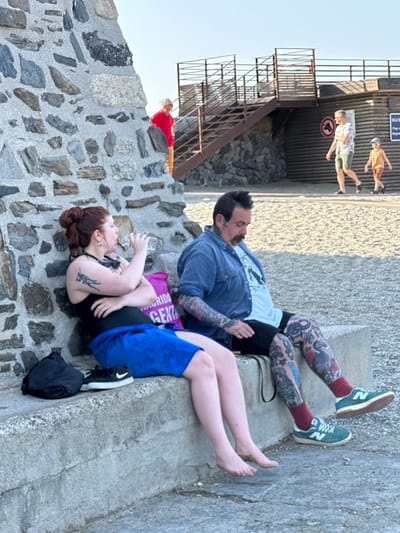 The image captures a scene on a paved area with a textured stone wall to the left. In the foreground, a woman with auburn hair tied up in a bun is seated, holding a clear plastic water bottle to her lips. She is wearing a black tank top and blue shorts, with bare feet resting on the ground. A black backpack and a pair of white sneakers with a black swoosh are visible next to her.

Beside her, a man with dark hair and a mustache is also seated. He is wearing a blue denim shirt over a white t-shirt and dark shorts. His legs are heavily tattooed. He is wearing dark red socks and green sneakers with a white "N" logo.

In the background, there are several people. A person in a red shirt and tan pants is standing on stairs leading up to a wooden structure with metal railings. Further in the background, a man and a young child are walking away from the camera on a sandy or pebbly surface. The man is wearing a patterned short-sleeved shirt and shorts, and the child is wearing a yellow shirt and brown shorts. The overall setting appears to be an outdoor public space, possibly near a beach or waterfront, with elements suggesting a tourist destination. The architectural style of the wooden structure and the presence of a sign with French text (likely "VILLE DE COLLIOURE" which would indicate Collioure, France) suggest the location is Collioure, France.