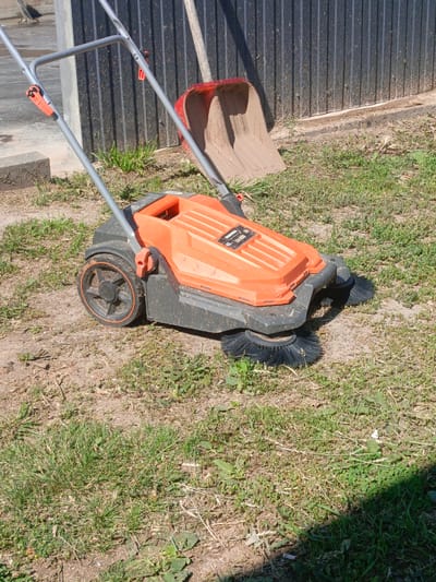 An orange and black mechanical sweeper sits on dry, patchy grass and dirt. The sweeper has two rotating brushes at the front and a handle with orange grips. Behind the sweeper, leaning against a dark corrugated metal fence, is a red shovel. The scene is outdoors, with a concrete structure visible to the left. The ground is mostly dry earth with sparse grass and weeds. The lighting suggests a sunny day.