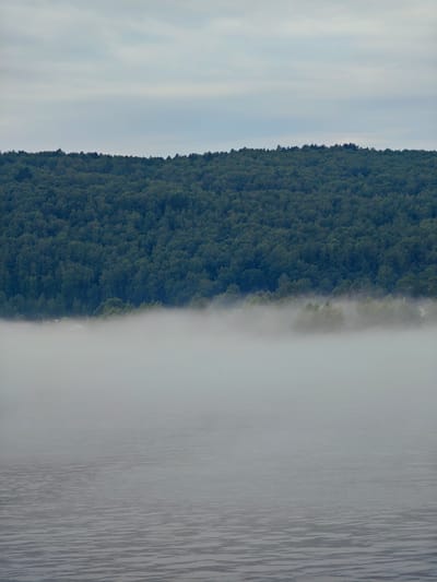 The image depicts a serene, misty landscape, likely captured from a body of water looking towards a densely forested shoreline. The foreground is dominated by the calm surface of the water, rendered in muted shades of gray with subtle ripples suggesting a gentle breeze. A thick layer of fog blankets the middle ground, obscuring the lower portions of the trees and creating an ethereal atmosphere. The fog appears to hover just above the water's surface, blending seamlessly with the hazy sky.
In the background, a vast, verdant forest stretches across the horizon. The trees are a deep, rich green, forming an unbroken canopy that rises towards the sky. The treeline is jagged, following the natural contours of the land. The sky above is overcast, with soft, diffused light filtering through the clouds.
The scene suggests a quiet, natural setting, possibly early morning or late evening, when fog is common. The presence of distant structures, faintly visible through the mist along the shoreline, hints at human habitation within the otherwise wild landscape, but these are too indistinct to identify. The overall impression is one of tranquility and natural beauty.
While the image itself does not contain specific landmarks to pinpoint the location in Atamanovo, Russia, such foggy conditions over bodies of water adjacent to forested areas are characteristic of many natural landscapes found in Russia during certain times of the year. The scene evokes a sense of peaceful isolation and the pervasive presence of nature.