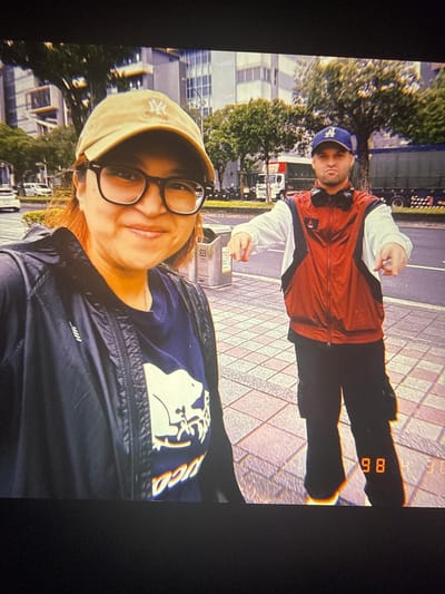 This is a photograph featuring two individuals in what appears to be an urban outdoor setting. The image is taken at an angle, suggesting a selfie or a candid shot.

In the foreground, a person wearing a light yellow baseball cap with a white logo and large dark-rimmed glasses is facing the camera. They have reddish-brown hair and are smiling. They are wearing a dark blue t-shirt with a graphic design on it and a dark jacket.

In the background, a second person is visible. They are wearing a blue baseball cap with a white logo, headphones around their neck, and a red, white, and black jacket. They are making a hand gesture and appear to be looking at the camera.

The setting includes a paved walkway with a diamond pattern, a street with vehicles, and buildings with windows. Trees with green foliage are also present. The lighting suggests it is daytime. There are some digital markings at the top right corner of the image that look like date or time stamps, possibly "86" and "85". Given the location context of Taipei, Taiwan, this could be a snapshot from their time there. The overall mood seems casual and cheerful.