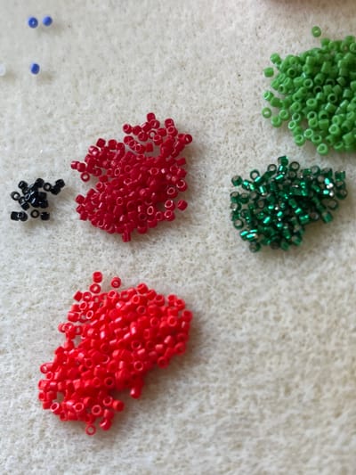 This image shows a close-up view of several piles of small seed beads scattered on a textured, light-colored surface that appears to be fabric or felt. The beads are of various colors, including bright green, shiny dark green, red, and a few scattered blue beads at the bottom left. There is also a small pile of black beads. The beads are cylindrical with a hole through the center, typical for beading or crafting. The lighting suggests an indoor setting, possibly a craft room or a table where someone is working on a project. The image is focused on the beads themselves, with the background slightly out of focus, highlighting the different textures and colors of the beads. There is no visible text. The scene suggests a hobby or craft activity is in progress or about to begin, with the beads laid out for selection. The focus is on the materials rather than a person or a completed project.