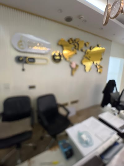 In the city of Karum, Nigeria, an office space is depicted. The wall is adorned with a gold-colored world map. In the foreground, two black chairs are visible, one of which is occupied by a person wearing dark clothing and carrying a tote bag. On a desk in the lower right corner of the image, there are several stacks of papers, with a blue object and a green document lying on top. The lighting in the room appears to be a mix of overhead lights and possibly natural light from a window, which is not fully visible.