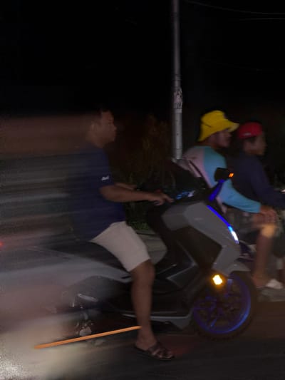 The image depicts a group of people gathered around a modern scooter or motorcycle at night. The scooter has a prominent blue-wheeled rim and a grey and black body. Several individuals are visible, some seated on or near the scooter, while others stand around it. One person wears a red cap and a yellow beanie, another a blue shirt and light-colored shorts. The scene appears to be outdoors, likely in a dimly lit area given the nighttime setting. The overall impression is that of a social gathering or a stop during a nighttime outing. There is no visible text in the image. The location context of Baan Plai Laem, Thailand, suggests this might be a local gathering or an event taking place in that specific region. The motion blur indicates movement, possibly during the capture of the photograph.