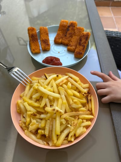 A child's hand is pointing towards a plate of fish fingers and a bowl of french fries on a glass table. The fish fingers are golden brown and arranged on a light blue plate with a dollop of ketchup. The french fries are golden yellow and piled high in a peach-colored bowl. A fork rests on the edge of the bowl, with some fries on it. The scene appears to be outdoors, possibly on a patio, with a glimpse of tiled flooring in the background. The child's hand is positioned to the right of the frame, with the index finger extended. The overall impression is a casual mealtime setting. The location is specified as Sant Feliu de Guíxols, Spain.
