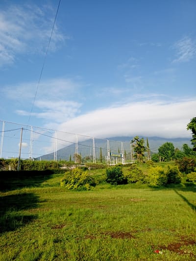 The image captures a scene of a grassy area in Thalang, Laos, set against a backdrop of a mountain partially obscured by a cloud formation. A white wire fence runs across the midground, behind which a row of bushes and trees borders the area. A sign is visible behind the fence. The sky is blue with scattered clouds. A power line is visible crossing the upper left portion of the frame.