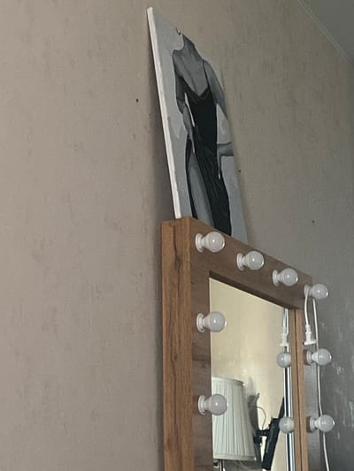 A framed painting of a woman in a black dress is leaning against a textured wall. Below the painting is a mirror with lights around its frame. The mirror reflects a lamp with a white pleated shade and some other objects. The scene appears to be indoors.