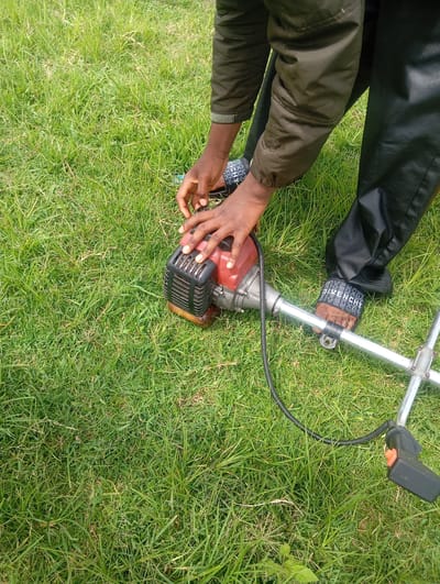A person, wearing a green jacket and black pants, is kneeling on a grassy area. Their hands are on a red and black string trimmer, which is partially visible. The person appears to be adjusting the trimmer. On their feet, they are wearing black slides with white lettering. The grass is green and appears to be freshly cut in some areas. The scene is outdoors, under what appears to be daylight. The person's focus is on the trimmer, suggesting they are preparing to use it or have just finished using it. The setting appears to be a natural environment, possibly a lawn or field, given the abundance of grass. There are no explicit cues about the time of day or weather, other than it being daylight. The location context provided is Gwafan, Nigeria. No discernable emotions are conveyed through the visible parts of the person. The visible text on the slides reads "GIVENCHY".
