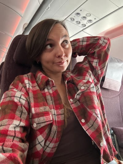 The image shows a woman lying in an airplane seat. She has brown hair and is wearing a brown shirt and a red and white plaid flannel shirt. Her head is tilted back, and she is looking up and to the left. The airplane window and overhead controls are visible in the upper left corner of the image. The background is a muted lavender color. It is not possible to determine the location as New Delhi, India, or any other location from the given information.