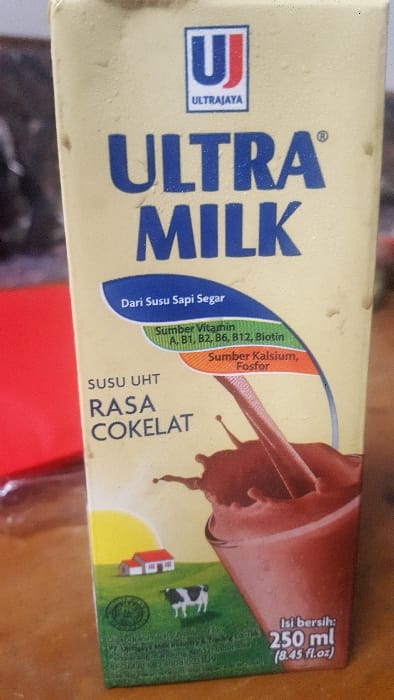 A beige-colored Ultra Milk carton, 250 ml (8.45 fl.oz.) in volume, stands upright, prominently centered. The carton displays the "ULTRA MILK" brand in large blue capital letters, with the "UJ" logo and "ULTRAJAYA" positioned above. The product is identified as "SUSU UHT RASA COKELAT" (UHT Milk Chocolate Flavor), made "Dari Susu Sapi Segar" (From Fresh Cow's Milk). Nutritional information highlights "Sumber Vitamin A, B1, B2, B6, B12, Biotin" and "Sumber Kalsium, Fosfor." Graphics on the label include a depiction of chocolate milk splashing into a glass, a rural scene with a single black and white cow, a small house with a red roof, and a yellow sun against a green landscape. A circular Halal certification logo is present. Micro-text at the base of the carton identifies the manufacturer as PT. Ultrajaya Milk Industry & Trading Co. Tbk., with the production facility located in Cimenyan, Bandung, Indonesia. The carton's surface exhibits visible condensation droplets. It rests on a brown, textured surface, possibly wood. To the left, a blurred red object or surface is partially visible, while the background remains indistinct and dark. No people are present. No specific actions or interactions are occurring within the visible scene.