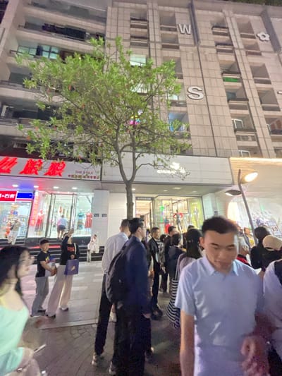 The image shows a busy city street in Hang Zhou Shi, China, located at coordinates 30.264 latitude and 120.156 longitude. A group of people are walking on the sidewalk in front of a large building. The building has a facade with the letters "W" and "S" prominently displayed. A large tree with green leaves grows in front of the building, and a street lamp casts a light over the scene. The building is adorned with windows, most of which have lights on inside, indicating that the scene takes place at night. The people are dressed in everyday clothing, and some are carrying shopping bags or other items. There are storefronts with signs in Chinese and English. Some of the storefronts are lit up with lights, and mannequins can be seen inside some of the windows. The people are walking in different directions, some looking at their phones. There is a sense of activity and energy in the air, indicating that this is a busy commercial district.