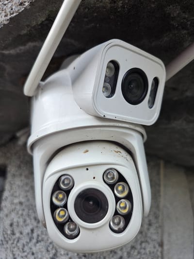 The image is a close-up, vertically oriented photograph of two white security cameras mounted outdoors. The cameras are positioned one above the other, with the upper camera slightly smaller and more rectangular, featuring a lens flanked by infrared illuminators. The lower camera is larger and more dome-shaped, with a central lens surrounded by eight infrared illuminators arranged in a circular pattern. Both cameras appear slightly dusty and weathered. The cameras are attached to a white antenna-like structure. The background consists of a textured, grey surface, possibly a wall or overhang, with some areas of rougher, stone-like texture. The image's orientation suggests it was taken from below, looking up at the cameras. There is no indication of human presence or activity within the frame. The location is identified as Marinka, Bulgaria.