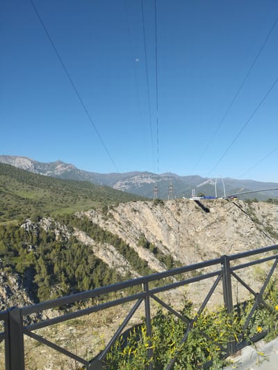 The image shows a wide shot of a canyon with a suspension bridge crossing it, located in the city of Myk, Uzbekistan. The sky is clear blue, with a faint crescent moon visible in the upper left quadrant. Several power lines stretch across the sky. The terrain is mountainous and rugged, with steep, rocky cliffs and hillsides covered in sparse vegetation, including trees and scrub.
In the mid-ground, the suspension bridge is prominent, connecting two sides of the canyon. The bridge has a modern design with white arching supports. On the cliff edge, near the bridge, there are several tall power pylons. The bridge area appears to be an attraction, with what looks like a sign with blue lettering visible on one of the platforms near the bridge.
In the foreground, a black metal railing is visible, with some green vegetation and small yellow flowers growing in front of it. The railing runs along the edge of a viewpoint, suggesting this is a tourist or scenic overlook. The overall scene depicts a natural landscape enhanced by man-made structures, likely a popular destination for its scenic views and adventure activities.