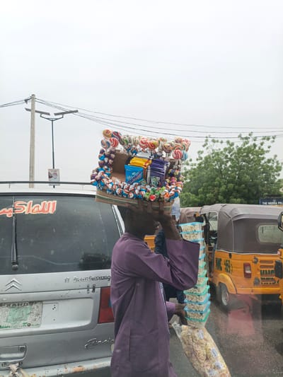 A street vendor in Hotoro, Nigeria, balances a tray of candy and snacks on his head, including lollipops, chocolates, and chewing gum in branded boxes. He is wearing a purple robe and carrying a stack of plastic containers filled with popcorn. The vendor is standing next to a silver Citroen car with Arabic script on the rear window and a heavily worn license plate. Behind him, there is a yellow auto rickshaw and a building that reads "THE NIGERIA POLICE." Overhead electrical wires are visible. The scene appears to be on a street. The sky is overcast.