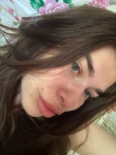 The image is a close-up, angled selfie of a young woman with long, dark brown hair that partially obscures her face. She has fair skin with some blemishes and freckles, and her eyes are a light grey-blue. Her lips are slightly parted and appear to be bare. The background is a floral-patterned fabric, likely bedding, with pink and green flowers. The lighting suggests it was taken indoors, possibly in a bedroom. The scene does not provide any information that would allow for the identification of a specific city or location within Мещёры, Russia.