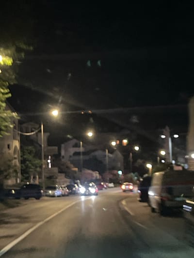 The image shows a nighttime street scene in Meljine, Montenegro. A road curves to the right, illuminated by streetlights and the headlights of vehicles. On the left side of the road, there are buildings with lights visible in some windows and trees with illuminated foliage. Several cars are parked or driving on the road, some with their headlights and taillights on. In the foreground, the road surface is visible, with a white line marking the edge. In the background, more buildings and streetlights can be seen, creating a sense of depth. The overall atmosphere is that of a quiet urban environment at night.