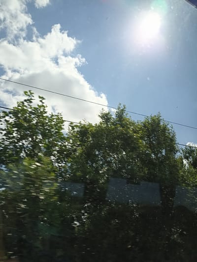 The image captures a view from inside a vehicle looking outwards. The sky is a clear blue, with several fluffy white clouds scattered across it. The sun is bright, positioned in the upper right quadrant of the image, casting a glare. In the mid-ground, a line of lush green trees is visible, their leaves appearing slightly blurred due to motion, suggesting the vehicle is in transit. Two dark, thin power lines stretch horizontally across the frame, intersecting the trees and sky. The lower portion of the image is dominated by the dark reflections on the vehicle's window, obscuring the ground and any potential details of the location within Clougherdiller, Ireland. No people are visible in the frame.