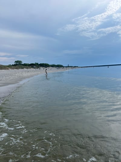 The image depicts a beach scene on a cloudy day. The foreground shows shallow water with gentle waves lapping at the shore, creating white foam. The beach itself is sandy, with dunes covered in vegetation lining the back of the shore. Numerous people are visible on the beach and in the water, appearing to be engaged in recreational activities. In the distance, a long structure, possibly a pier or breakwater, extends into the sea. The sky is overcast with scattered clouds. The scene is consistent with a public beach in Baltiysk, Russia, known for its coastal locations.