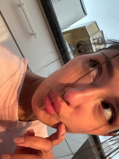 A close-up, selfie-style photograph captures a woman indoors. She has light skin, dark hair with strands across her face, and is wearing a pale pink shirt. Her expression includes pursed lips and a gaze directed upwards and to the left. Her right index finger touches her cheek.
In the background are household appliances, including a cream-colored device and a white appliance, situated on a dark countertop above white cabinets with horizontal bar handles.
There is no indication of the location being Miljevci, Montenegro in this image.