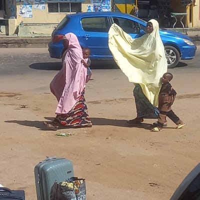 The image depicts a daytime street scene in Sardauna, Nigeria, featuring several individuals and a parked blue car on an unpaved road. The sunlight is bright, casting distinct shadows, suggesting it's either mid-morning or late afternoon.
In the foreground, on the left, a woman is seen standing near the blue hatchback car. She wears a light yellow, flowing head covering that drapes over her body. Her face is visible, and she looks towards the viewer. To her right, a young child stands, dressed in a brown long-sleeved top, dark trousers, and light yellow shoes. The child appears to be interacting with or standing very close to the woman in yellow.
Further to the right, another woman is present, distinguished by her pink head covering and a pink garment worn over a patterned skirt or dress. She is carrying a baby on her back, who is partially visible and wearing a striped top.
On the far right side of the frame, two pieces of luggage are visible on the ground. One is a light blue/grey hard-shell suitcase, and on top of it rests a black bag with a colorful pattern, where faint text like "SHOP" can be discerned. Another dark suitcase is partially visible below the light blue one.
In the background, behind the car, there is a building with light-colored walls and windows. Several posters or flyers are affixed to the building's facade, though their text is illegible. The ground throughout the scene appears to be dirt or sand, indicating an unpaved road or shoulder. The overall impression is one of people gathered or waiting by the roadside, possibly preparing for travel.