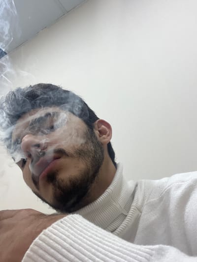 A young adult male, characterized by dark hair, a full beard, and a mustache, is depicted from a low-angle perspective, exhaling a visible plume of smoke from his mouth and nose. He is dressed in a white turtleneck sweater, with a portion of his right hand discernible in the lower-left foreground. The immediate environment is an interior space, featuring a plain white wall and a faint ceiling junction line visible in the upper-left background. This scene is located in Al-Mazza Municipality, Syria.
