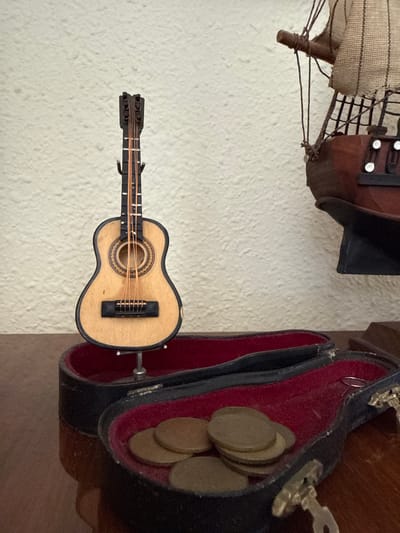 A photograph shows a miniature guitar lying next to a partially open black case lined with red velvet, containing several coins. Above these items is a portion of a model ship. The objects are set against a plain, light-colored wall with a textured surface. The exact location in Holon, Israel cannot be determined from the image.