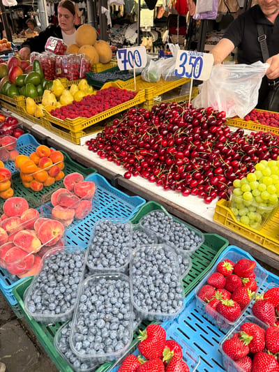 A bustling outdoor market stall in Vračar Urban Municipality, Serbia, displays an abundant selection of fresh fruits. In the foreground, various containers overflow with vibrant strawberries, their red hues contrasting with the deep blue of blueberries. To the right, a crate of bright green grapes adds another pop of color. Behind these, large piles of ripe cherries are showcased, with a smaller assortment of apricots visible further back. The stall also features rows of peaches and yellow pears, alongside avocados and what appear to be red currants in plastic containers. Melons are stacked on top, and a vendor, a woman with dark hair wearing a dark jacket over a white shirt, is seen arranging the fruit. Another vendor, a man with dark hair and a black shirt, is visible on the right, handling a plastic bag. Price signs, one reading "700 KILO" and another "SLATKA 350 KILO," are displayed, indicating the market's commerce. The scene is captured under natural lighting, highlighting the freshness and variety of the produce.