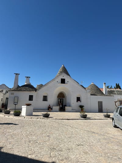 The image depicts a sunny daytime scene in Alberobello, Italy, showcasing the iconic Trulli houses. In the foreground, a cobblestone street is visible, with shadows cast by unseen objects. Several Trulli houses, characterized by their conical roofs and whitewashed walls, line the street. Some of these structures have chimneys protruding from their roofs. Planters with small green plants are placed near the houses, and one planter sits atop a white pedestal. Two women are seated on a step in front of one of the Trulli houses. To the right, the rear of a silver car is partially visible. The sky is clear and blue.