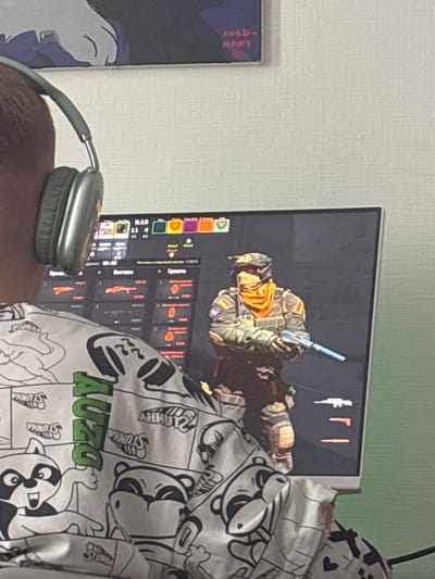 A person with gray headphones is in front of a computer monitor. The monitor displays what appears to be a video game. The person is wearing a white hoodie with cartoon-like raccoon designs and the word "AUZO" written on it in green. The location appears to be a room with light-colored walls. Alyumino, Russia cannot be confirmed as location.