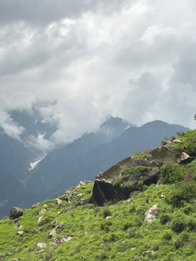 The image shows a mountainous landscape with lush green vegetation covering the slopes in the foreground. Large rocks are scattered across the grassy hillside, with one prominent rock formation in the mid-right of the frame covered in moss. In the background, multiple layers of dark, misty mountains rise up, with patches of snow visible on some of the higher peaks. The sky is overcast with heavy, grey clouds, obscuring the sun and creating a diffused light. The scene depicts a natural, remote environment, characteristic of the Himalayas near Bahang, India. There are no people or animals visible in the image.