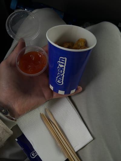 A person is holding a cup of chicken nuggets with a side of sauce in their left hand. A white napkin and chopsticks are next to the hand. The cup is blue and white and says "Check In" on the side. The background is gray and appears to be someone's lap. The sauce is a reddish-orange color. There is no information to support the location in the city of Getamej, Armenia.