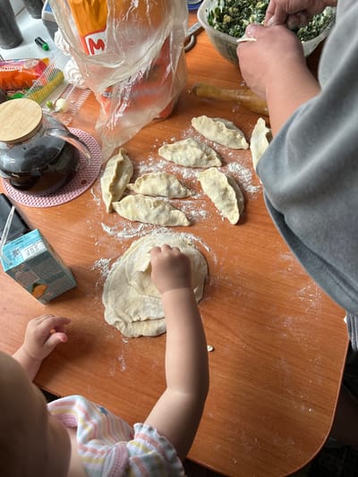 In a home setting, likely in the city of Azinsky, Russia, a wooden table is laden with ingredients and partially prepared food. A baby, wearing a striped top, is seated at the table, reaching out to a pile of dough with their right hand. Next to the baby is a blue carton of milk. In the center of the table, several uncooked dumplings are laid out, dusted with flour. To the right, an adult's hands are visible, engaged in the process of making dumplings, with a bowl of green filling and a rolling pin nearby. In the background, a large plastic bag, a glass teapot with dark liquid, and other food items are scattered across the table, suggesting ongoing food preparation. The lighting indicates it is daytime, with a light source from the left.