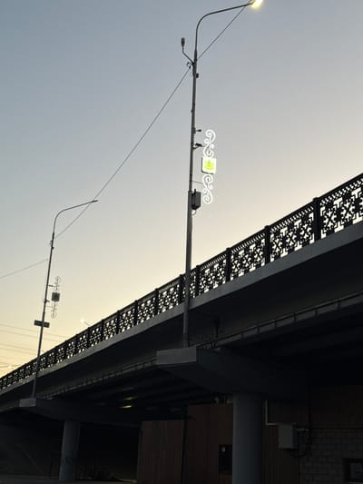 This is a media file, likely a photograph, showing a view looking up towards an elevated structure, possibly a bridge or overpass, against a pale, overcast sky. The perspective is tilted, making it appear as if the scene is sideways.
The main subjects are structural elements. A prominent object is a streetlight with a single light fixture, casting a warm, yellow glow. Extending from this streetlight is a horizontal arm supporting a decorative element with a yellow and white illuminated sign. The sign has a stylized floral or crest-like design. Wires are visible, strung between various points on the structure and the streetlight.
Underneath the elevated structure, decorative panels with intricate, dark cut-out patterns are visible. These panels are affixed to the underside of the structure, creating a rhythmic visual effect. Further to the right, parts of a building facade are visible, constructed with wooden planks and brickwork. A small, dark rectangular opening, possibly a window, is set into the wooden section.
The time of day appears to be late afternoon or early evening, suggested by the soft, diffused light and the illuminated signs. The weather seems to be cloudy or hazy, contributing to the muted tones of the sky. There are no people clearly visible. The overall mood is calm and somewhat industrial due to the visible infrastructure. No discernible text is present within the image. The location context provided is Novyi Chirkei, Russia.