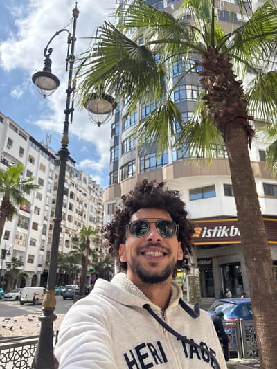 An upward-angled selfie captures a male individual smiling in a bustling urban environment identified as Tanger, Morocco, specifically near an "Istikbal" storefront. The subject, positioned centrally, has dark curly hair, a beard, and wears reflective sunglasses, a light-colored hooded sweatshirt with "HERITAGE" partially legible, and a dark lanyard around his neck.
Behind and to the left of the subject, an ornate black lamppost with two classical streetlights rises vertically. To the subject's right, a prominent palm tree trunk extends upwards, obscuring part of a modern building with multiple reflective windows. Further in the background, to the left, multiple multi-story residential or commercial buildings with balconies are visible.
At street level, below the lamppost and to the left of the subject, a street features several parked and moving vehicles, including a white commercial van and a blue car. A group of pigeons congregates on the ground. A decorative metal railing is visible in the bottom left corner. To the right, behind the palm tree, the "Istikbal" store's prominent brown and orange signage is visible, along with parts of its storefront and blurred figures on the sidewalk. The sky is partly cloudy with patches of blue.