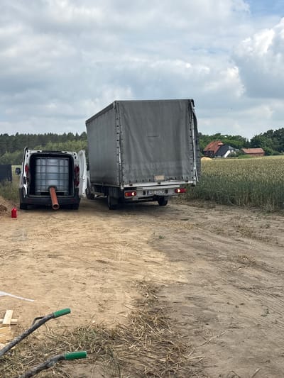 In Opatów, Poland, on a dirt road leading to a field of wheat, a white van with its rear doors open is parked. Inside the van, a large plastic water tank is visible, with a brown pipe extending from its base. Next to the van, a small red plastic container lies on the ground. Behind the van, a large truck with a grey tarp-covered trailer is parked, its license plate reading "PKE 2JC9". In the background, a dense forest lines the horizon, and beyond that, a few houses with red roofs are nestled among trees. The sky is overcast with scattered clouds. In the foreground, two dark hoses with green ends lie on the dry, sandy ground, along with some scattered straw and a few wooden planks.