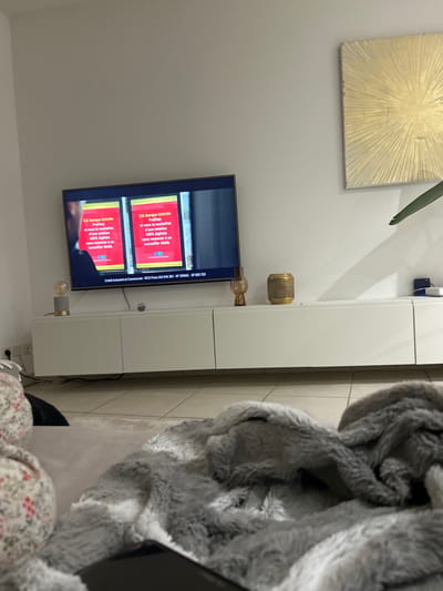 The image shows a modern living room with a television displaying a commercial advertisement for CIC Banque Hybride. The ad emphasizes the benefits of their hybrid banking service, highlighting features like personalization and a 100% digital experience. The setting includes a wall-mounted TV above a white cabinet and abstract art on the wall. A fluffy gray blanket and a floral-patterned garment add a touch of domesticity to the space.