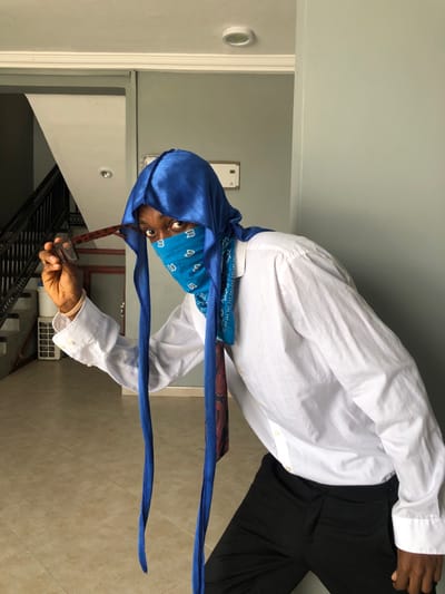 An individual with dark skin is depicted indoors in Gbawe, Ghana, leaning slightly forward and looking directly at the camera. The subject's head is covered by a blue silky durag, which drapes over the head and extends into two long ties. A light blue bandana with a white paisley pattern covers the lower half of the face, obscuring the nose, mouth, and chin. A dark blue tie with a red paisley pattern is visible hanging from the neck area, beneath the bandana and durag. The individual wears a white long-sleeved button-up shirt and dark (likely black) trousers. The right hand (viewer's left) is raised, holding a pair of folded brown tortoiseshell-patterned eyeglasses by one arm.

The environment features light-colored tiled flooring and light grey walls. In the background on the left, a dark-railed staircase ascends, with a white rectangular object visible near its base. On the wall in the right-center background, a white rectangular panel is mounted. A circular white ceiling light fixture is visible overhead.