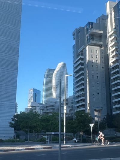 The image shows a street scene in Givatayim, Israel, characterized by modern high-rise buildings under a clear blue sky. In the foreground, a person is riding a bicycle from right to left across the frame. The cyclist, wearing a black tank top and shorts, is facing away from the camera. The bicycle is dark-colored with a light-colored basket or bag attached to the handlebars.

The middle ground features several trees and some lower-rise structures, including a bus stop shelter. Behind the trees, the dominant elements are the tall buildings. On the far left, a very tall, grey, rectangular skyscraper with horizontal lines is visible. To its right are two distinctive, curvilinear white skyscrapers with horizontal banding. Further to the right, a grey, stepped skyscraper with numerous balconies is prominent. Another blue-toned glass skyscraper is also visible in the distance.

The street in the foreground has a striped curb, indicating a pedestrian crossing or a traffic boundary. A lamppost is positioned near the center of the frame, and other street signage and poles are scattered throughout the scene. The lighting suggests it is daytime, with the sun casting shadows and highlighting the architecture.