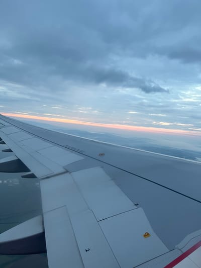 The image presents an aerial view from the window of an aircraft, likely operating in the airspace near Lozen, Bulgaria, during either sunrise or sunset. The dominant feature is the aircraft's wing, which extends diagonally across the lower and middle sections of the frame, from the lower left to the mid-right.
The wing is predominantly white or light grey, displaying visible panel lines, rivets, and several distinct control surfaces on its trailing edge. Towards the center of the wing's upper surface, two prominent yellow, U-shaped latch or tie-down mechanisms are visible. Further along the wing, a black warning inscription states "NO STEP" and "DO NOT WALK OUTSIDE THIS AREA." A red stripe, part of the aircraft's livery, is partially visible on the fuselage in the lower right corner, adjacent to the wing root. The number "2" is marked on a panel towards the lower left portion of the wing.
Above the wing, the sky is largely covered by layered, broken clouds, exhibiting shades of grey and light blue, with some brighter, diffused light areas. Along the distant horizon, a narrow, horizontal band of soft pastel colors – primarily pink, orange, and pale yellow – indicates the low position of the sun, characteristic of either dawn or dusk. Beneath this colored horizon, the landscape appears hazy and indistinct, presenting a vast expanse of muted blue-grey, suggesting distant landforms or a body of water obscured by atmospheric mist. The immediate foreground below the left side of the wing shows a darker, less defined area, possibly a body of water or obscured terrain. No individuals are visible in the scene.
