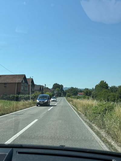 The image depicts a view from inside a vehicle traveling on a paved road. The road has white lane markings and is bordered by a grassy shoulder and trees on the right side. In the distance, there are houses with red tiled roofs and lush green foliage. Two vehicles are visible on the road ahead. The one in front is a white car, and following it is a blue van. The sky is clear and blue, suggesting a sunny day. The scene appears to be a rural or suburban setting in Mali Zvornik Municipality, Serbia, likely during daylight hours given the bright sky. The perspective indicates the photo was taken while the vehicle was in motion. No people are clearly discernible within the frame, although the driver of the blue van is partially visible. The time of day is likely midday or early afternoon due to the bright sunlight and clear sky. There is text on the license plate of the blue van, but it is not legible.