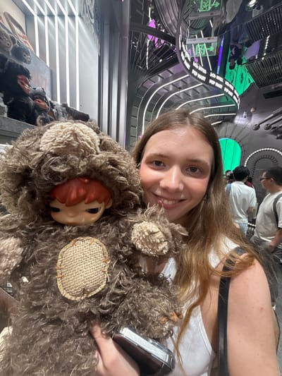 A fair-skinned young woman with long, light brown hair smiles at the camera while holding a stuffed animal. The toy is a brown, furry creature with a beige patch and a doll-like face with orange hair. They appear to be inside a futuristic-looking building with curved metal walls and ceilings, illuminated by white and purple lights. In the background, other people can be seen walking. Based on the merchandise visible in the background, the image was taken at the Zootopia-themed land inside the Shanghai Disneyland Resort, located in Shanghai, China.