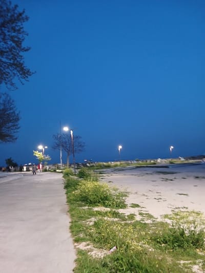 The image shows a paved walkway along a waterfront in İstanbul, Türkiye, at coordinates 40.956, 28.816. The walkway is flanked on the right by a sandy area overgrown with small green plants and on the left by a low concrete wall and a tree. The sky is a deep blue, and the only light comes from streetlights, which are visible in the distance. A few people are walking along the walkway, their silhouettes visible against the blue of the evening sky. The scene is tranquil and serene, with a sense of peace and quiet.