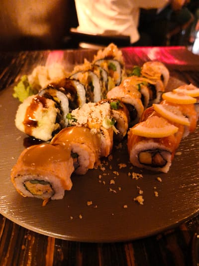 A plate of sushi rolls is presented on a textured, dark plate. The sushi appears to be a variety of rolls, some with a fried coating and others with what looks like raw fish or salmon on top, garnished with thin slices of lemon. Drizzled sauces, possibly teriyaki or spicy mayo, are visible on several of the rolls.
In the background, out of focus, a person is partially visible, appearing to be in a dining setting. The lighting suggests an indoor environment, possibly a restaurant, with a warm ambiance. The overall impression is of a prepared meal ready for consumption.
The image's context is Be'er Sheva, Israel. No text is visible in the image.