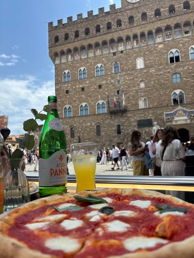 Here is a description of the image:
The image shows a table setting with a pizza, a bottle of Acqua Panna water, and a glass of yellow liquid, possibly lemonade or another beverage. The pizza appears to be a Margherita, with tomato sauce, mozzarella, and basil leaves. The background reveals the Palazzo Vecchio in Florence, Italy. People are visible in the piazza, including tourists and locals, walking around and interacting. The weather seems sunny with a clear blue sky. The overall scene captures a typical dining experience in Florence, with the iconic Palazzo Vecchio providing a recognizable backdrop.