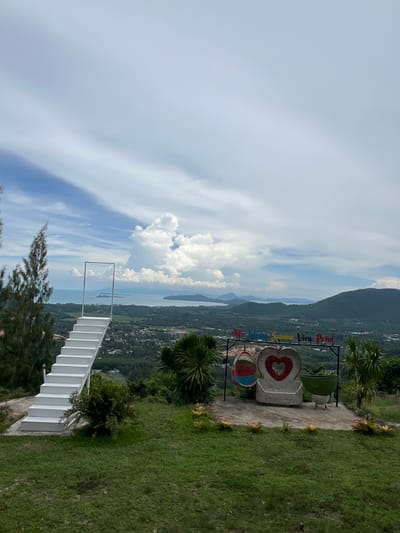 This is a full shot of a viewpoint in Baan Thurian, Thailand, offering a panoramic view of the coast and lush green hills. In the foreground, a white staircase with railings leads up to a framed opening that overlooks the expansive landscape. To the right of the staircase, there is a decorative setup featuring a heart-shaped stone bench, two swings, and a large green pot. The background is dominated by a vast sky filled with dramatic clouds, with the sea and distant islands visible on the horizon. The landscape below consists of rolling hills covered in dense vegetation and scattered buildings. The area around the viewpoint is covered in grass, with a few trees and plants adding to the natural scenery. The weather appears to be partly cloudy, with patches of blue sky visible between the clouds.