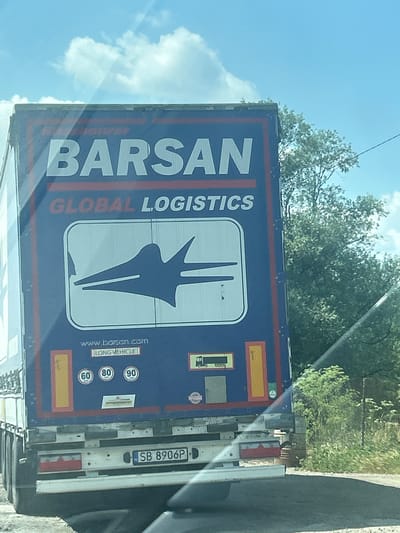 A blue trailer truck with "BARSAN GLOBAL LOGISTICS" written on the back is traveling on a road. The trailer has a dark blue logo on the back that appears to be an abstract representation of a bird in flight. There are speed limit stickers indicating 60, 80, and 90. The truck's license plate is "SB 8906P". The truck is seen from behind, and is on a road with trees and vegetation on the right side. The sky is blue with some clouds. The image was captured in the vicinity of Momin Prohod, Bulgaria.
